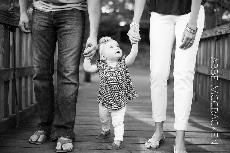 one year old holding parents hands, learning to walk looks to mom for reassurance