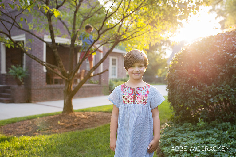 Summer lifestyle portrait of a girl with strong backlighting, rim lighting, brother climbing tree in background