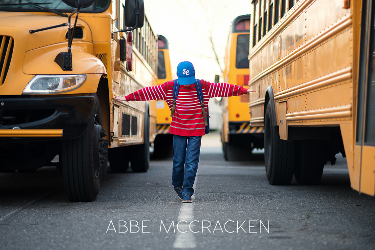 young school boy walking on a parking line in between school buses