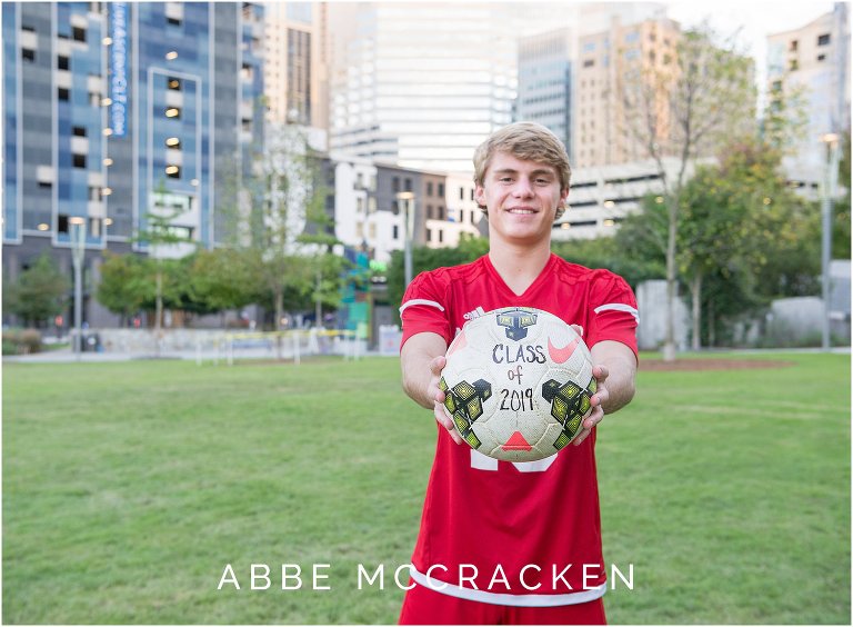 Senior boy wearing his Charlotte Catholic soccer uniform and holding a "class of 2019" soccer ball