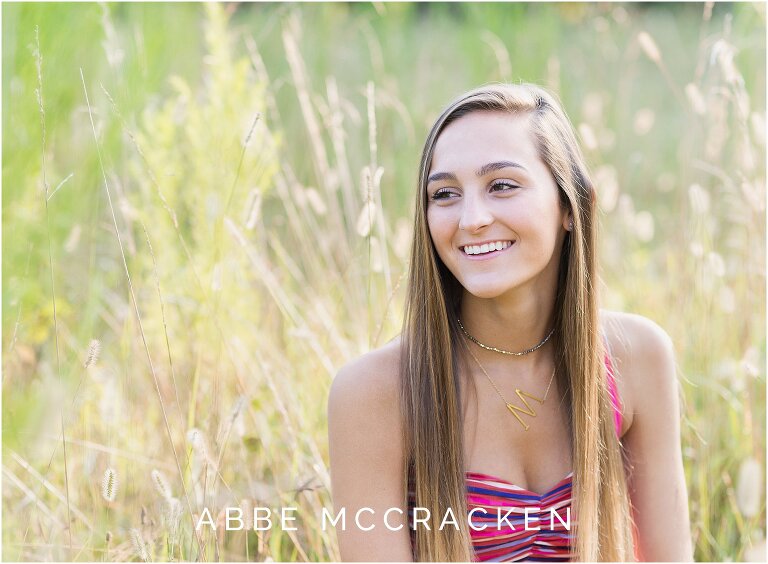 candid picture of a high school senior girl in colorful striped dress sitting in tall wheat grasses