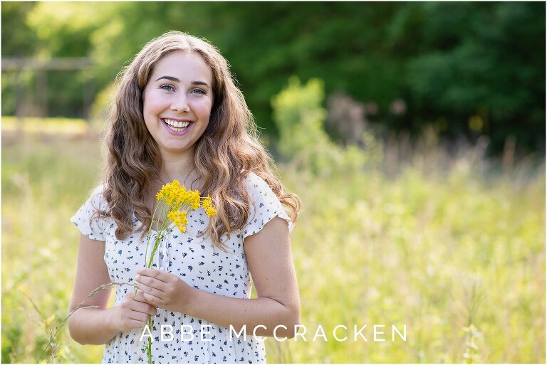 senior girl with brown curly hair poses with a yellow flower