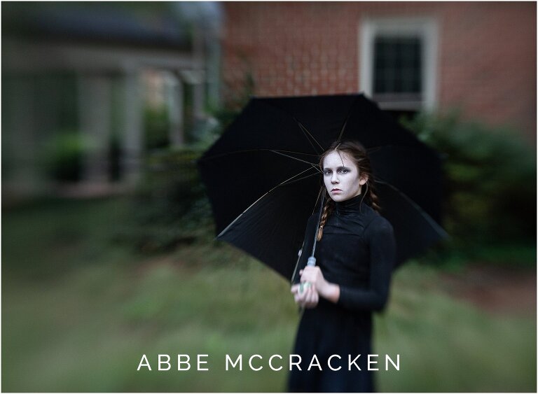 Halloween portrait of a ghost girl in all black and full white makeup, holding an umbrella and staring blankly into camera. Lensbaby used for extra blur and mystery.