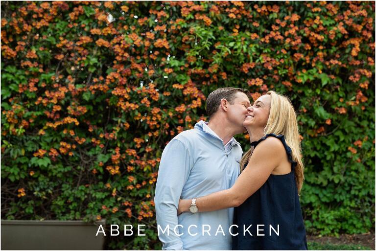 husband kissing his wife's cheek in Uptown Charlotte, wall of colorful orange flowers behind them
