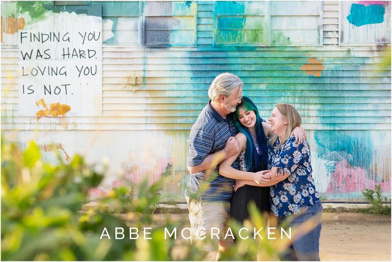 Candid senior portrait of parents hugging daughter, layers of greenery in foreground and colorful mural in background