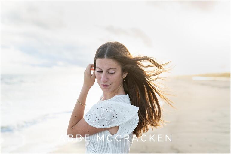 Gorgeous candid senior portrait of a girl on the beach with dark hair blowing in the wind, sun is backlighting her hair