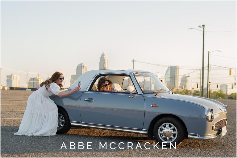 Image of a high school senior sitting in a vintage car while his mother takes a picture of him. The skyline of Charlotte NC is in the background.