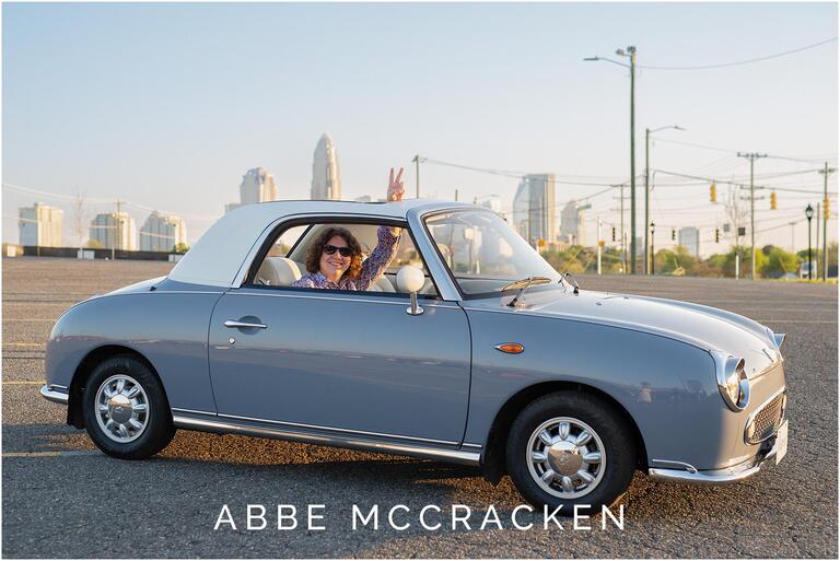 Senior portrait of a boy sitting in a vintage car with the Charlotte skyline in the background. Taken from the parking lot at Camp North End.