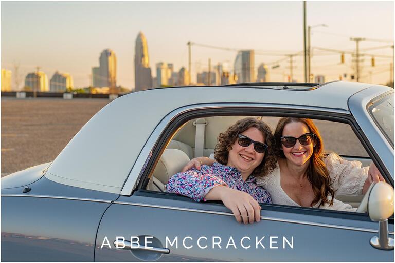 Portrait of a high school senior with his mom, both sitting in a vintage car with the Charlotte skyline in the background