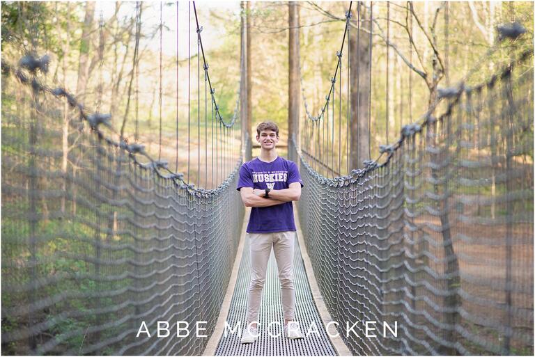 High school senior boy standing on the swinging bridge at Anne Springs Close Greenway, wearing college t-shirt and khaki pants