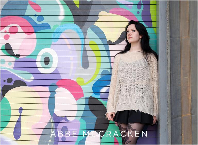 Senior portrait of a girl with porcelain skin and long dark hair standing against a colorful mural at Camp North End in Charlotte, NC