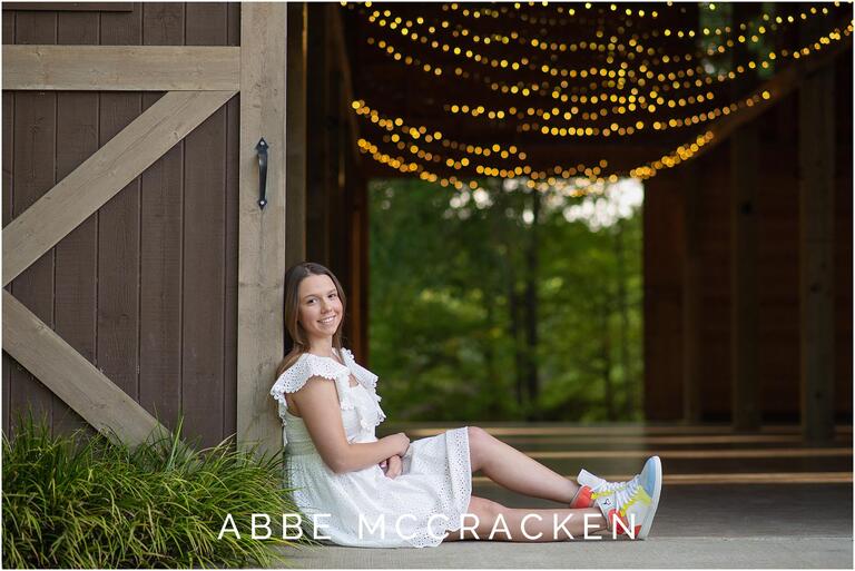 Charlotte senior pictures - girl sitting against barn door with twinkling lights above