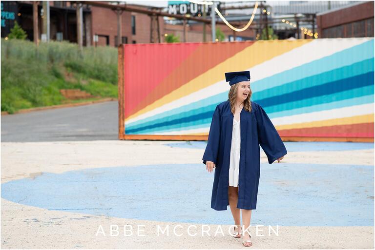 Senior girl wears cap and gown in front of rainbow wall art and strung lights at Camp North End for graduation photos