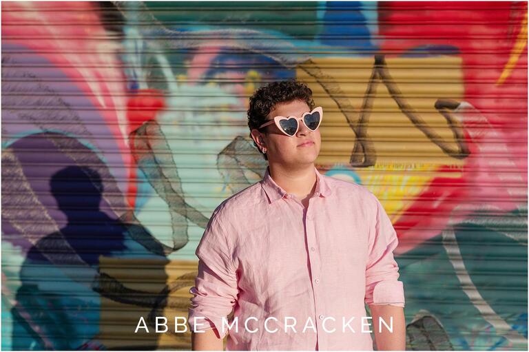 Senior portrait of a guy in pink shirt and pink heart sunglasses in front of a colorful Camp North End mural, heavy shadow on background