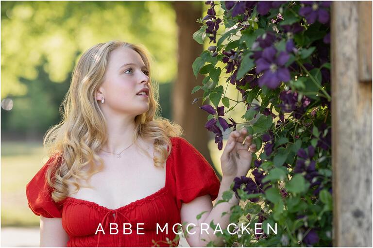 Blonde, fair skinned senior girl in red dress looking at purple climbing flowers