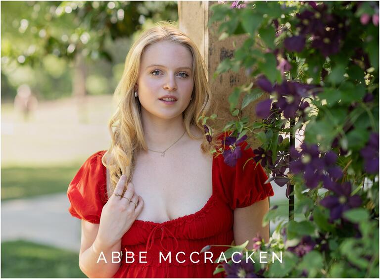 Blonde, fair skinned senior girl in red dress standing beside purple climbing flowers