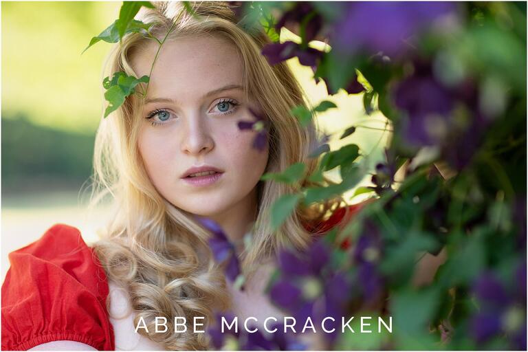 Stunning portrait of a fair-skinned high school senior in red dress peeking through the purple clematis in Charlotte's Independence Park