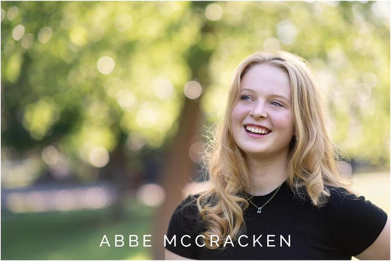 Candid image of a high school senior laughing as she looks to the sky, bokeh in the background as light comes through the trees