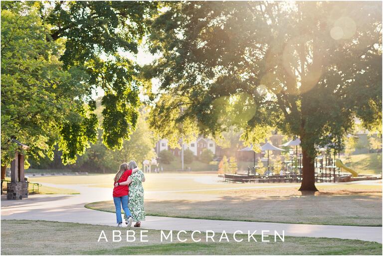 Mother and daughter walking through Charlotte's Independence Park after daughter's senior portrait session, lens flare coming in top right of image from sun