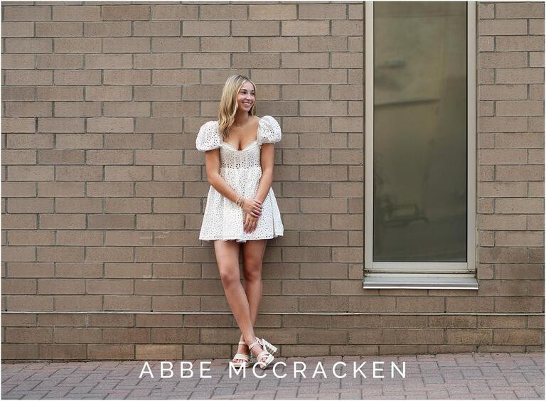 Senior portrait of a girl in white dress standing on a tan brick wall