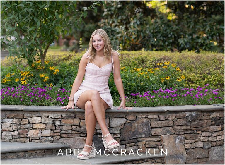 Summer senior portrait of a beautiful blonde in short, tweed dress sitting on stonework in The Green, surrounded by flowers