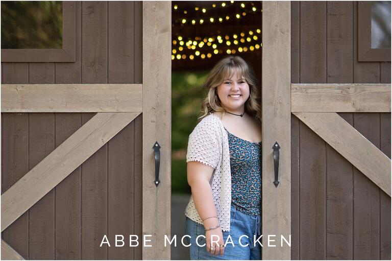 Image of a high school senior standing between barn door, twinkling white lights in the background