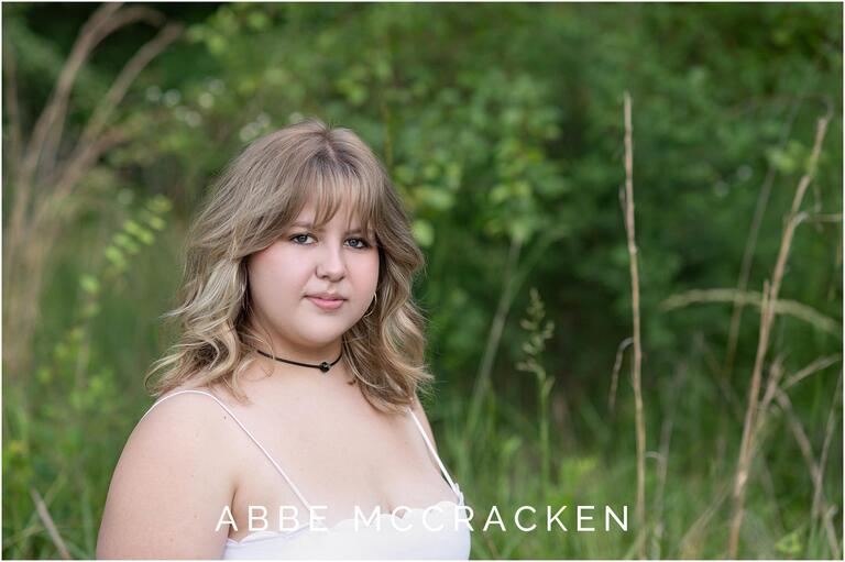 Senior portrait of a blonde in white sundress, background is green fields and forest